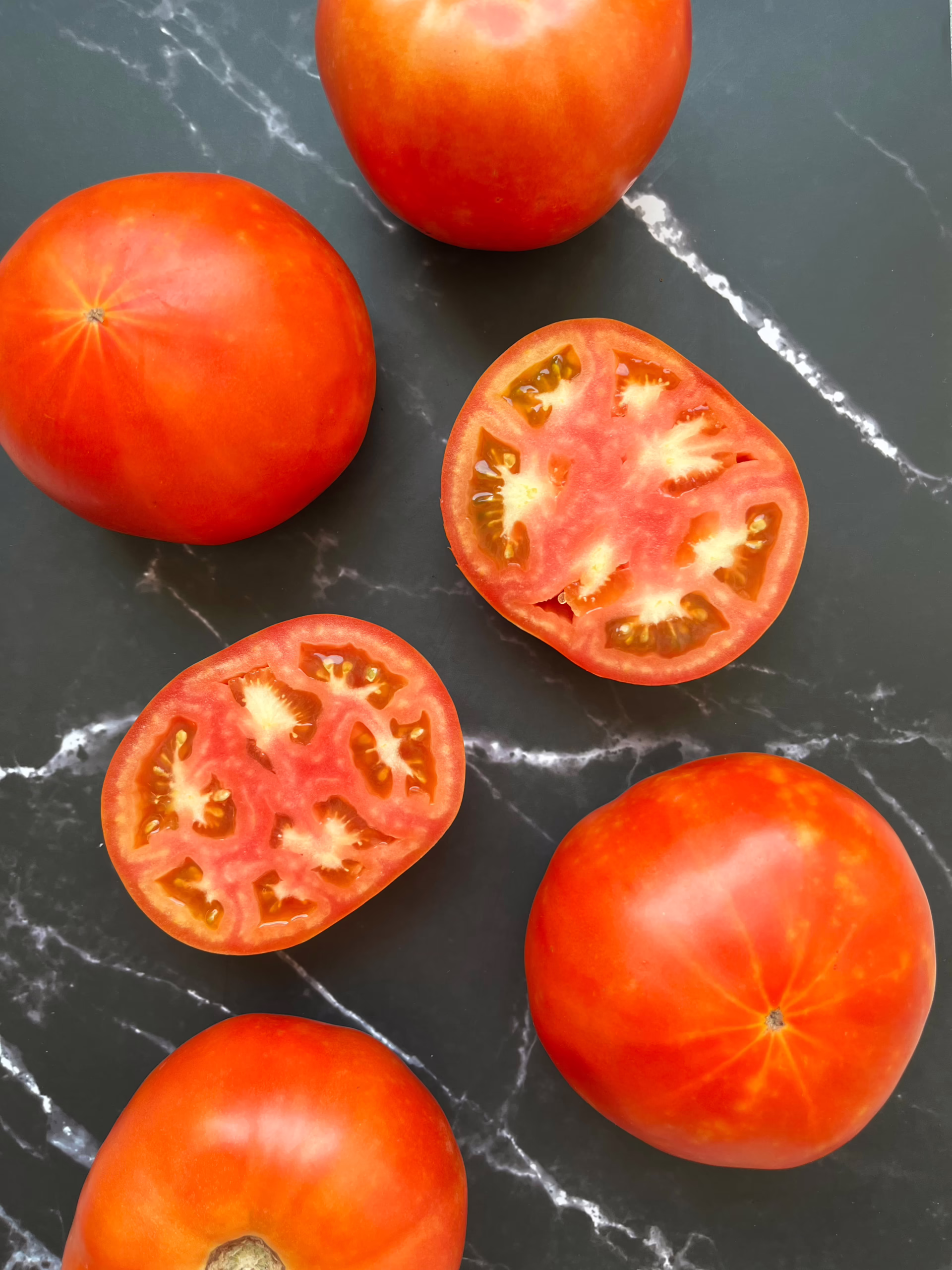 Five red tomatoes, three whole and two cut in half, are arranged on a black marble surface. Beefsteak tomatoes, tomato size chart