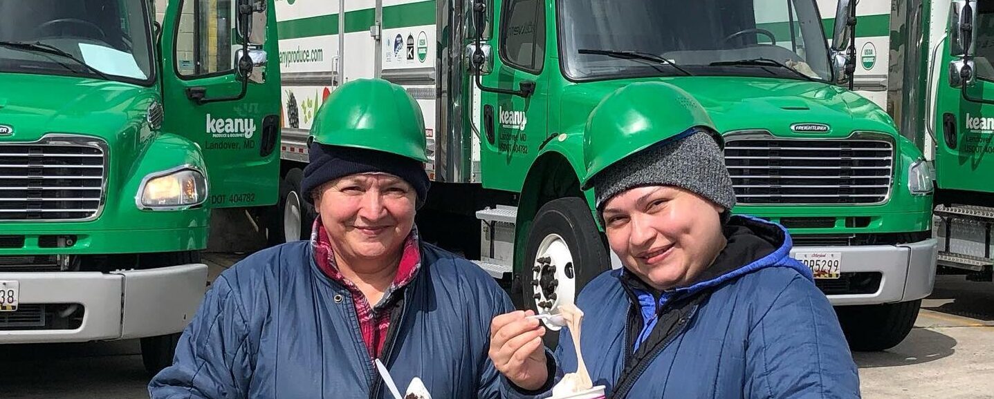 Two people in green hard hats and blue jackets stand in front of green trucks, savoring ice cream as they discuss jobs in fresh produce.