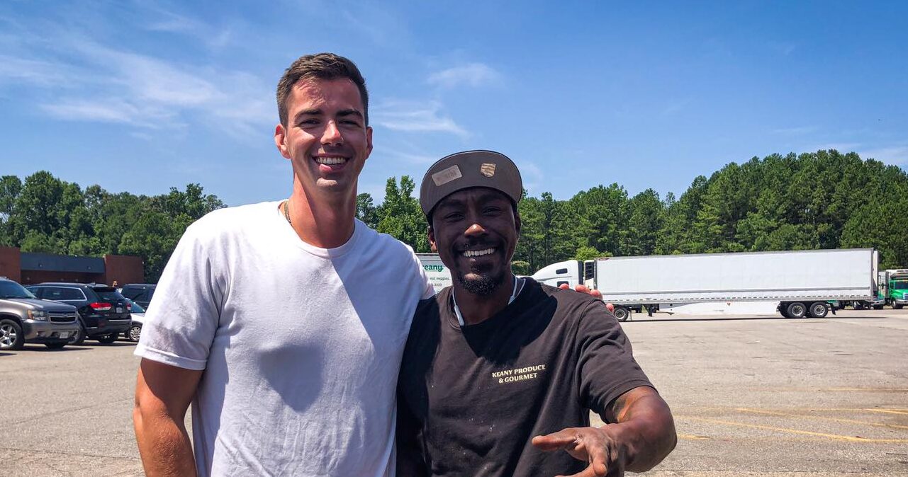 Two men smiling in a parking lot, one wearing a white t-shirt and the other in a dark shirt and cap, perhaps discussing their produce careers, with trucks and trees in the background.