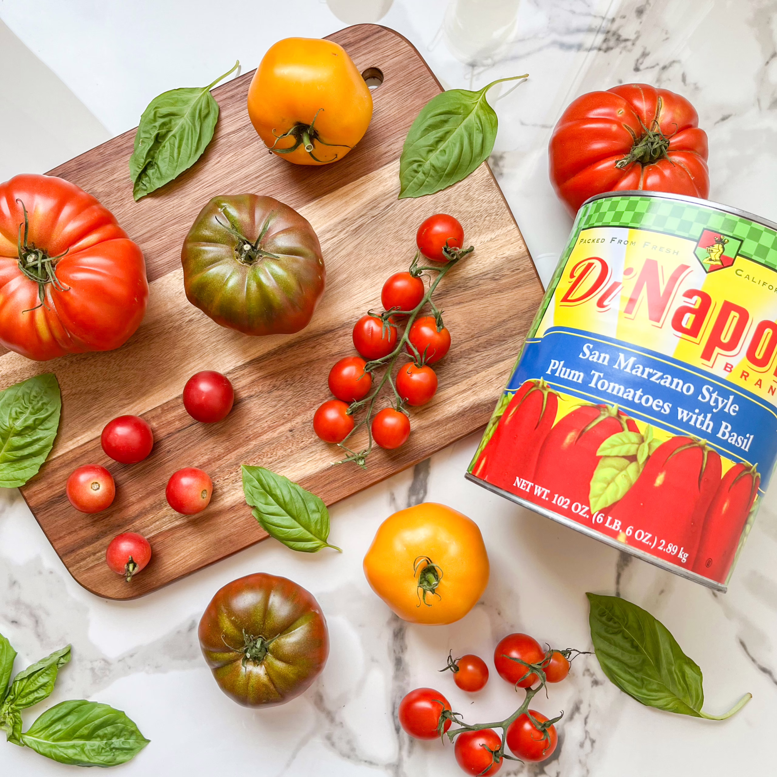 Tomato Size Chart - Know Your Tomatoes A variety of fresh tomatoes and basil leaves on a marble surface, with a large can of San Marzano style plum tomatoes and a wooden cutting board. Tomato size chart